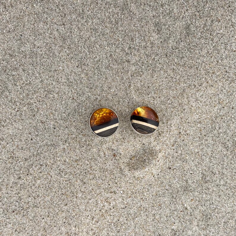 May include: A pair of amber and black striped earrings with silver settings. The earrings are on a sandy surface.