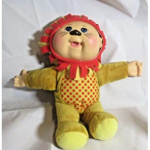 May include: A vintage doll dressed as a lion. The doll has a round face with blue eyes, a smiling expression, and a red and yellow lion's mane. The doll's body is yellow with a red polka-dot pattern on the front.
