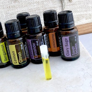 Op de afbeelding: Een verzameling van zeven amberkleurige glazen flessen met zwarte doppen, die elk 15 ml etherische olie bevatten. De flessen zijn gelabeld met de namen van de oliën: Bergamot, Citroen, Citron, Lavendel, Wierook, Salie en Clary Sage. De flessen zijn in een rij op een wit oppervlak gerangschikt. Een klein, doorzichtig glazen flesje met een gele vloeistof bevindt zich op de voorgrond.