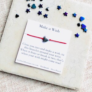 Op de afbeelding: Rood koord armband met een zilveren hart bedel. De kaart staat "Make a Wish" en bevat instructies over hoe je een wens kunt doen met het armband. De kaart staat ook "LilyDailyBoutique.etsy.com Handmade Silver Plated".