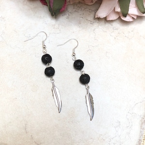 Könnte beinhalten: Ein Paar silberne Ohrhänger mit drei schwarzen Lavasteinen und einem silbernen Federschmuck.