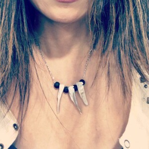 May include: A woman with long brown hair and a necklace with black and white beads. The necklace has three white pendant charms that look like teeth.