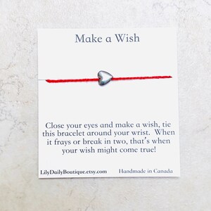 Op de afbeelding: Rood koord armband met een zilveren hart bedel. De kaart staat "Make a Wish" en bevat instructies om het armband om je pols te binden en een wens te doen. Wanneer het armband rafelt of breekt, kan je wens uitkomen. De kaart bevat ook de website LilyDailyBoutique.etsy.com en vermeldt "Handmade in Canada".