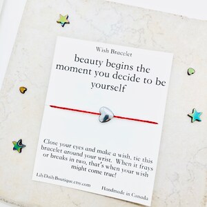 May include: Red string wish bracelet with a silver heart charm. The card reads "Wish Bracelet, beauty begins the moment you decide to be yourself. Close your eyes and make a wish, tie this bracelet around your wrist. When it frays or breaks in two, that's when your wish might come true! LilyDailyBoutique.etsy.com Handmade in Canada"