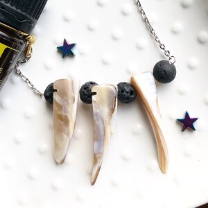 May include: A silver chain necklace with three white shell pendants and three black lava rock beads. The necklace is on a white plate with a small bottle of essential oil labeled "dōTERRA Helichrysum Helichrysum italicum" and two iridescent stars.