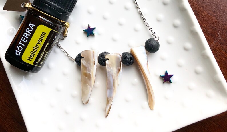 May include: A silver chain necklace with three white shell pendants and three black lava rock beads. The necklace is on a white plate with a small bottle of essential oil labeled "dōTERRA Helichrysum Helichrysum italicum" and two iridescent stars.