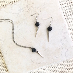 Puede incluir: Un conjunto de joyas que incluye un collar y pendientes. El collar tiene una cadena plateada con una cuenta de lava negra y un colgante de punta plateada. Los pendientes tienen ganchos plateados, cuentas de lava negra y dijes de punta plateada. El conjunto se muestra sobre una superficie blanca y beige.