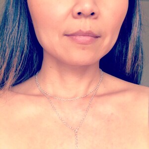 May include: A woman wearing a black tank top with a silver chain necklace with a blue eye charm pendant.