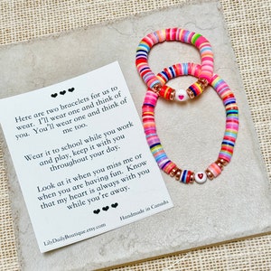 May include: Two colorful bracelets with heart charms. The bracelets are made of colorful beads and have a message card that reads "Here are two bracelets for us to wear. I'll wear one and think of you. You'll wear one and think of me too. Wear it to school while you work and play, keep it with you throughout your day. Look at it when you miss me or when you are having fun. Know that my heart is always with you while you're away. LilyDailyBoutique.etsy.com Handmade in Canada"