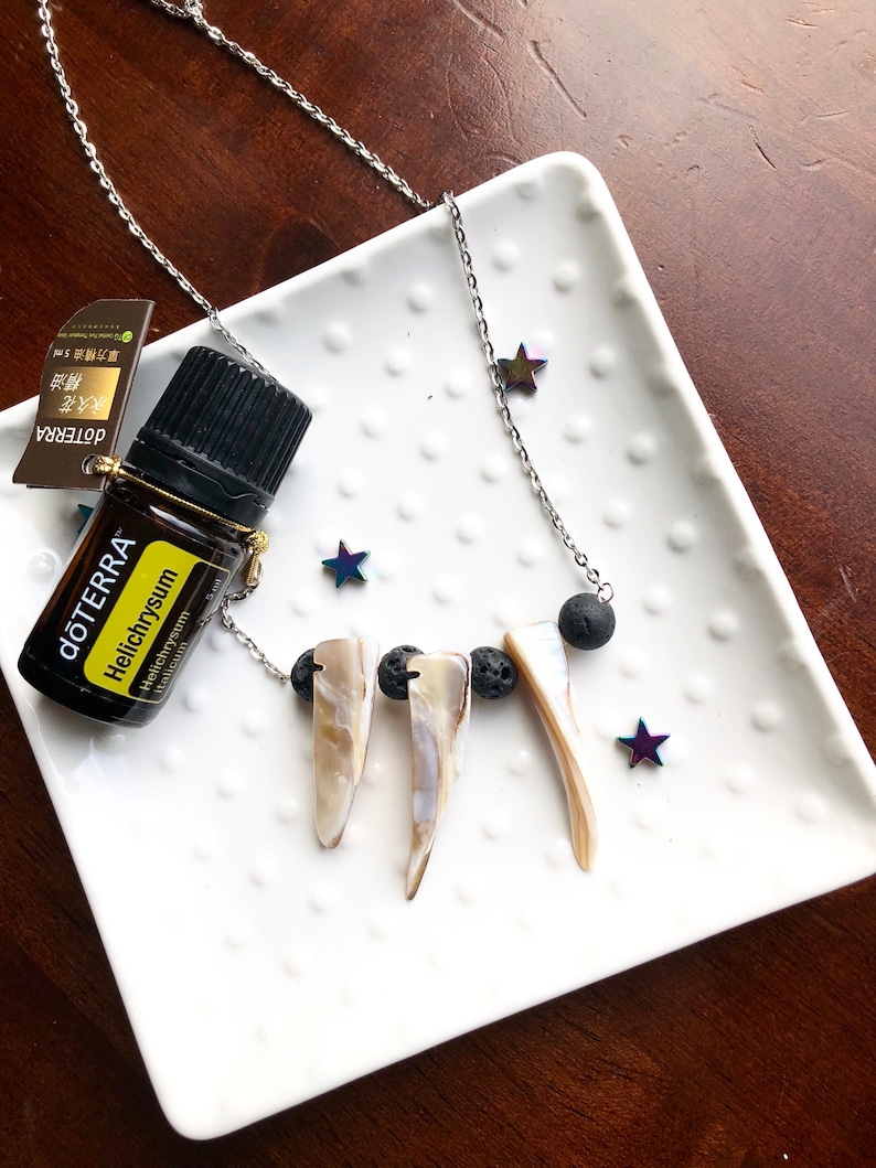 May include: A silver chain necklace with three white shell pendants and black lava rock beads. A small bottle of essential oil labeled "dōTERRA&reg; Helichrysum" is also in the image.