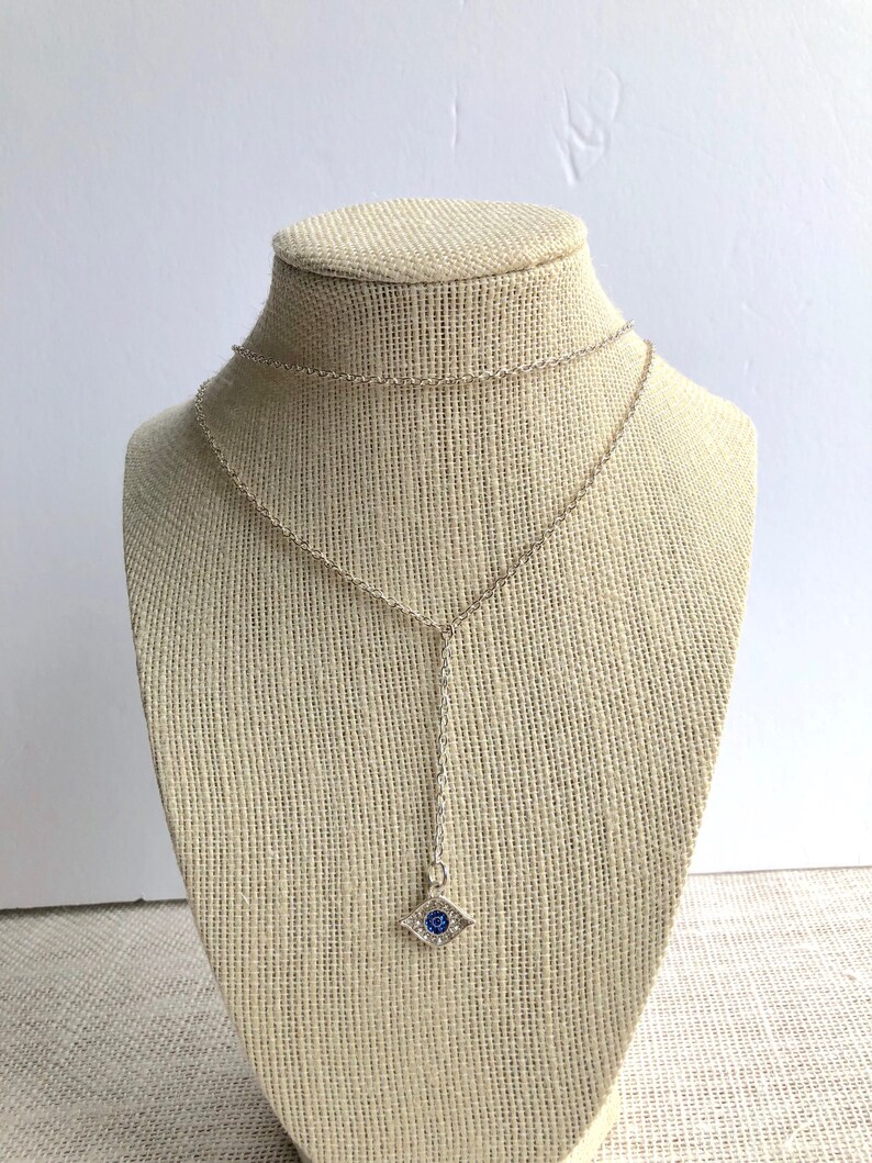 May include: A silver chain necklace with a small blue evil eye pendant. The pendant is surrounded by clear rhinestones.