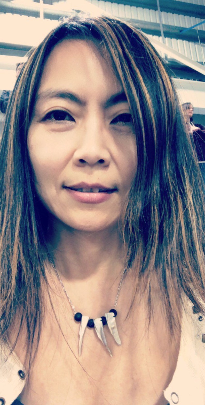 May include: A woman with long brown hair and a necklace with black and white beads. The necklace has three white pendant charms that look like teeth.