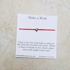 Op de afbeelding: Rood koord armband met een zilveren hart bedel. De kaart staat "Make a Wish" en bevat instructies om het armband om je pols te binden en een wens te doen. Wanneer het armband rafelt of breekt, kan je wens uitkomen. De kaart bevat ook de website LilyDailyBoutique.etsy.com en vermeldt "Handmade in Canada".