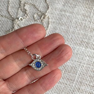 May include: A silver chain necklace with a blue and white evil eye pendant. The pendant is covered in small clear rhinestones.