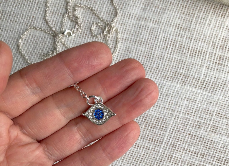 May include: A silver chain necklace with a blue and white evil eye pendant. The pendant is covered in small clear rhinestones.