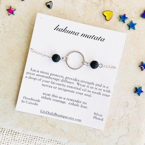 Puede incluir: Un brazalete plateado con un dije circular y dos cuentas de lava negra. El brazalete está sobre una tarjeta blanca con el texto "hakuna matata" y "lleva esto como un recordatorio para: inhalar coraje. exhalar miedo." La tarjeta también incluye el texto "Hecho a mano en Canadá" y "LilyDailyBoutique.etsy.com".