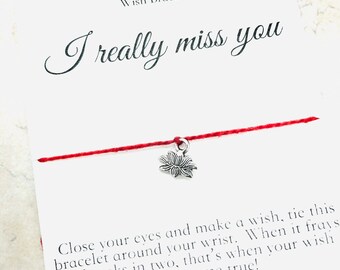 May include: Red string wish bracelet with a silver flower charm. The card reads "I really miss you" and includes instructions on how to make a wish. The card also says "Handmade in Canada" and includes a website address.