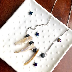 May include: A silver chain necklace with three iridescent shell pendants and black lava rock beads. The necklace is displayed on a white plate with iridescent star-shaped accents.