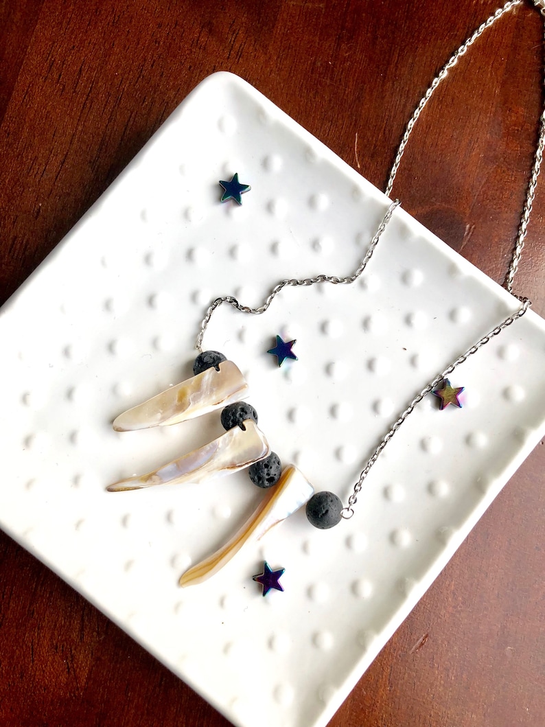 May include: A silver chain necklace with three iridescent shell pendants and black lava rock beads. The necklace is displayed on a white plate with iridescent star-shaped accents.