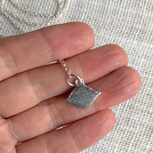 May include: A silver chain necklace with a small, silver pendant shaped like a pillow. The pendant is textured and has a slightly rounded shape.