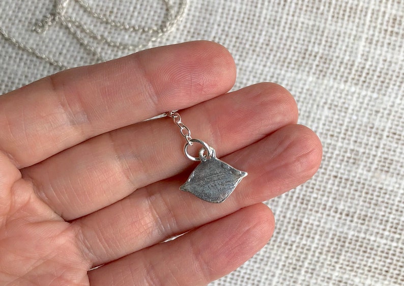 May include: A silver chain necklace with a small, silver pendant shaped like a pillow. The pendant is textured and has a slightly rounded shape.