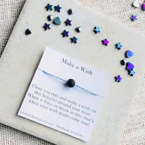 Może przedstawiać: Bransoletka z niebieskiego sznurka z czarną kulą lawy. Bransoletka jest przywiązana do kartki z napisem "Make a Wish" i zawiera instrukcje, jak złożyć życzenie. Na kartce jest również napisane "Close you eyes and make a wish, tie this bracelet around your wrist. When it frays or break in two, that's when your wish might come true!" oraz "LilyDailyBoutique.etsy.com Handmade in Canada."