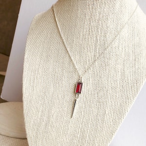 Puede incluir: Un collar de plata con un colgante en forma de daga adornado con una piedra preciosa roja. El colgante está en una cadena de plata.