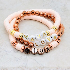 Op de afbeelding: Drie gelaagde armbanden met roze en gouden kralen. De bovenste laag spelt "MAMA" in witte letters, de middelste laag spelt "LOVE" in witte letters en de onderste laag spelt "DIOR" in gouden letters.