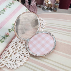 May include: An open, vintage-style compact mirror with a round mirror and a pink gingham patterned interior. The compact has a distressed, metallic frame and is set on a white lace doily. The background features a floral-patterned book and striped fabric.