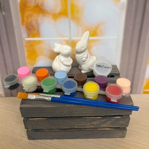 May include: Two small white ceramic rabbit figurines sit atop a dark gray wooden crate. In front are two blue-handled paintbrushes and a strip of twelve small plastic paint pots in various colors, plus one separate pot labeled Sealant.