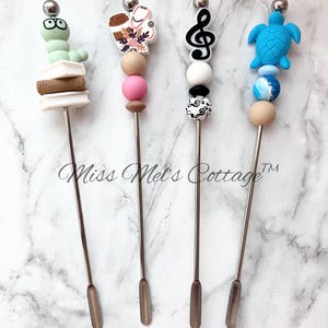 May include: Four stainless steel stir sticks with decorative tops. The tops feature a green worm with glasses, a coffee cup and stethoscope, a musical treble clef, and a blue turtle. The text "Miss Mel's Cottage" is in the image.