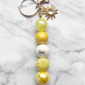 May include: A yellow and white beaded keychain with a gold sun charm and a white bead that says "You are my sunshine".