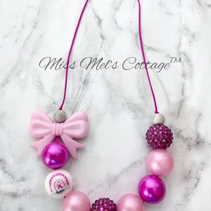 May include: A pink beaded necklace with a bow, round beads, and a pendant with a rainbow and the word "hope". The necklace has a pink cord and silver accents. The necklace is on a white marble surface.