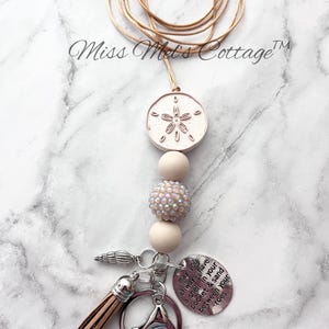 May include: A beige lanyard with a sand dollar pendant, iridescent bead, and a tassel. The lanyard includes a keychain with a charm that reads "may you always have a shell in your pocket & sand between your toes."