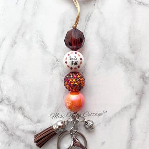 May include: A beaded lanyard with a brown tassel and key rings. The lanyard features a series of decorative beads in shades of brown, orange, and white. The white bead has the text "Hello Fall" printed on it.