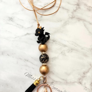 May include: A lanyard featuring a black dragon charm with gold horns, two metallic gold beads, one black and gold speckled bead, and two rhinestone spacers. The design includes a black tassel and a rose gold lobster clasp on a tan cord.
