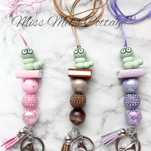 May include: Three colorful beaded lanyards with a book-reading worm charm, each with a tassel and a silver key ring. The lanyards are pink, brown, and purple.