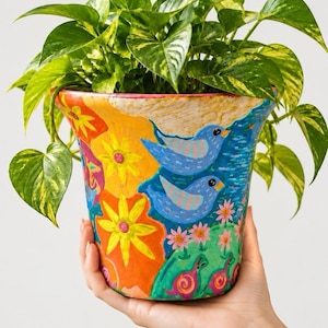Pode incluir: Um vaso de cerâmica pintado à mão com ilustrações vibrantes de dois pássaros azuis, flores amarelas e laranjas brilhantes, flores cor-de-rosa e pequenos caracóis cor-de-rosa. Uma planta jiboia verde com folhas em forma de coração cresce no vaso.