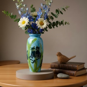 May include: A light blue ceramic vase featuring hand-painted black cats and green foliage, filled with a bouquet of white daisies, blue delphinium, and eucalyptus leaves. The vase has a tapered neck and a smooth, glossy finish.
