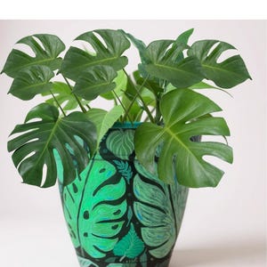 以下が含まれることがあります： 大きな鮮やかな緑色の葉を持つ、鉢植えのモンステラ。植物は、黒い背景と緑のモンステラの葉の手描きのデザインが施された装飾的な鉢に入っています。鉢にはターコイズの縁があります。