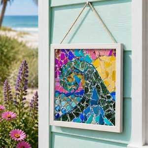 May include: A square wall art piece featuring a mosaic design of a swirling ocean wave. The artwork is encased in a white wooden frame and hangs from a jute rope. The mosaic uses colourful glass pieces in shades of blue, green, yellow, and pink.