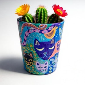 Puede incluir: Maceta de cerámica pintada a mano con un diseño colorido de gatos, pájaros y formas abstractas. Contiene tres pequeños cactus, cada uno con una flor amarilla o roja. Una pieza de decoración única, de unos 10 cm de altura.