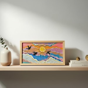 Könnte beinhalten: Ein gerahmtes Kunstwerk zeigt einen lebendigen Sonnenuntergang mit fliegenden Vögeln. Das Gemälde zeigt eine gelbe Sonne, einen rosa-orangenen Himmel und blaue Wolken. Das Kunstwerk wird auf einem Holzregal mit dekorativen Gegenständen ausgestellt.