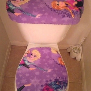 May include: Purple toilet seat cover and lid with a white border. The cover features a design of three cartoon girls with snowflakes. The lid features a design of two cartoon girls with snowflakes.