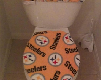 May include: A toilet seat and lid with a Pittsburgh Steelers themed fabric cover. The cover is orange with black and white Steelers logos and the word "Steelers" repeated throughout.