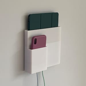 Puede incluir: Organizador blanco de pared con dos compartimentos. El compartimento superior contiene una funda de tableta verde oscuro y el compartimento inferior contiene una funda de teléfono rosa. Dos cables cuelgan de la parte inferior del organizador.