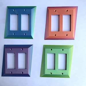 Puede incluir: Cuatro placas de pared dobles en diferentes colores iridiscentes: verde azulado, naranja, morado y verde. Cada placa tiene dos aberturas rectangulares para interruptores de luz o tomas de corriente.