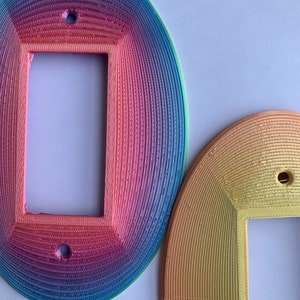 以下が含まれることがあります： 虹色のグラデーションデザインが施された、カラフルな丸い装飾的なコンセントカバーが2つ。カバーには、電気コンセント用の長方形の開口部があります。