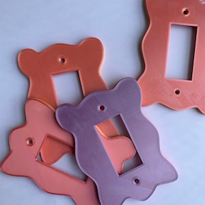 May include: Three decorative light switch covers in pink and orange with a wavy, organic shape. The covers are made of plastic and have a matte finish.