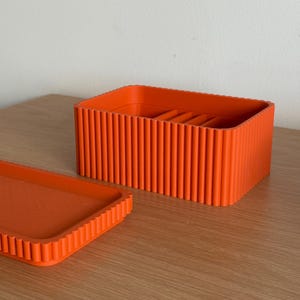 Pode incluir: Uma caixa de armazenamento retangular laranja com uma tampa correspondente. A caixa e a tampa têm um design canelado. A caixa está aberta, revelando divisórias internas. Os itens estão em uma superfície de madeira.
